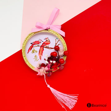 A round preserved flower ornament with a tassel, featuring a watercolor-painted bird design on fabric, accompanied by cherry blossoms and red berries, finished with a pink ribbon bow.